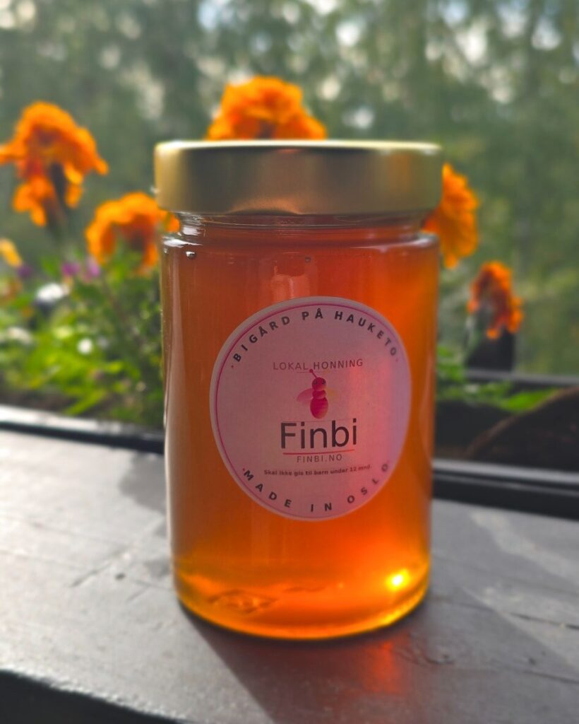 Et glass flytende Finbi-honning med rosa etikett, fotografert ute blant blomster